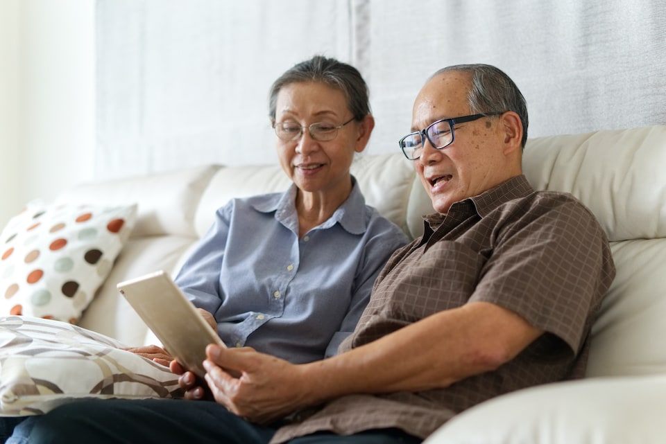 Senior couple sitting on sofa in home playing tablet and relaxing together. They are smiling and enjoy to spend their time together with happiness. Happy retirement life concept; downsizing tips