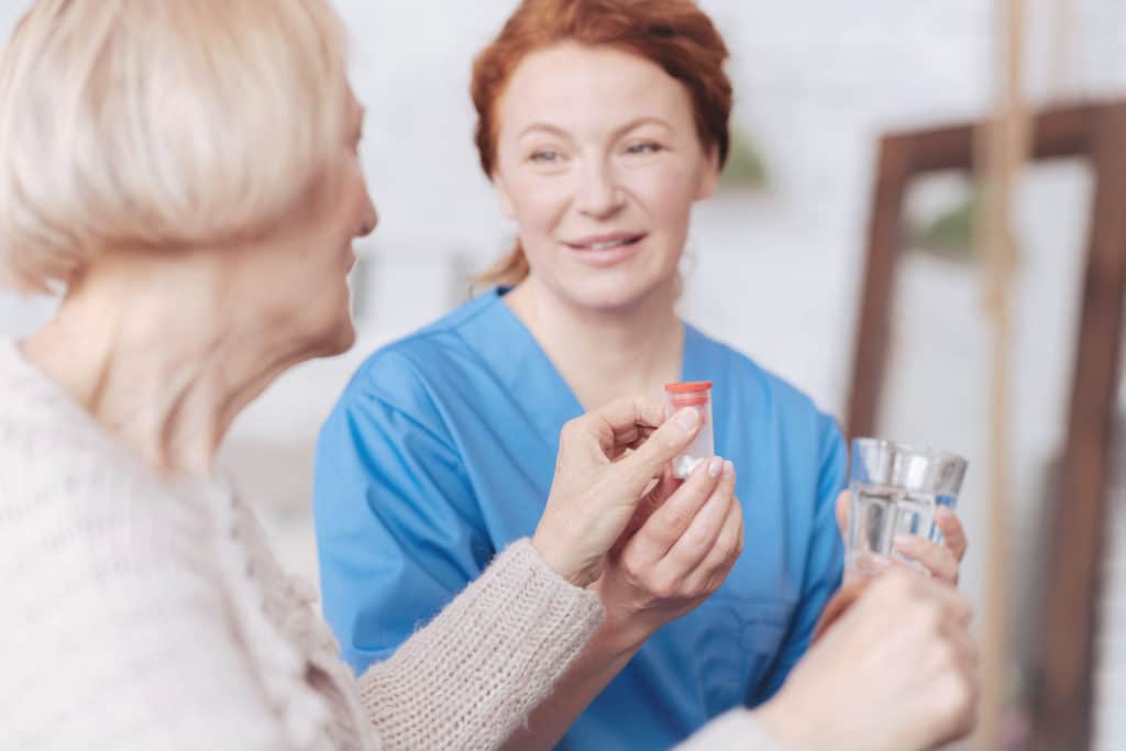 Daily routine. Selective focus on a hand of a senior lady sitting on the edge of her bed and taking a bottle with her prescription while talking to her mature caregiver.