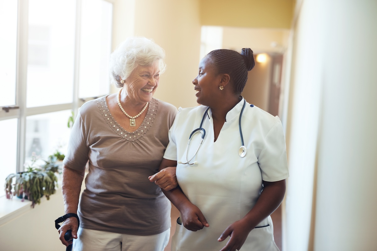 Senior woman walking in the nursing home supported by a caregiver, both smiling