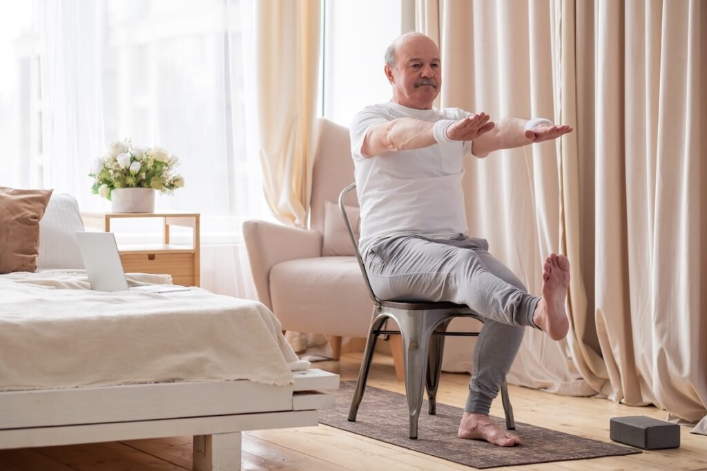 Elderly man practicing yoga asana or sport exercise for legs and hands using chair. 