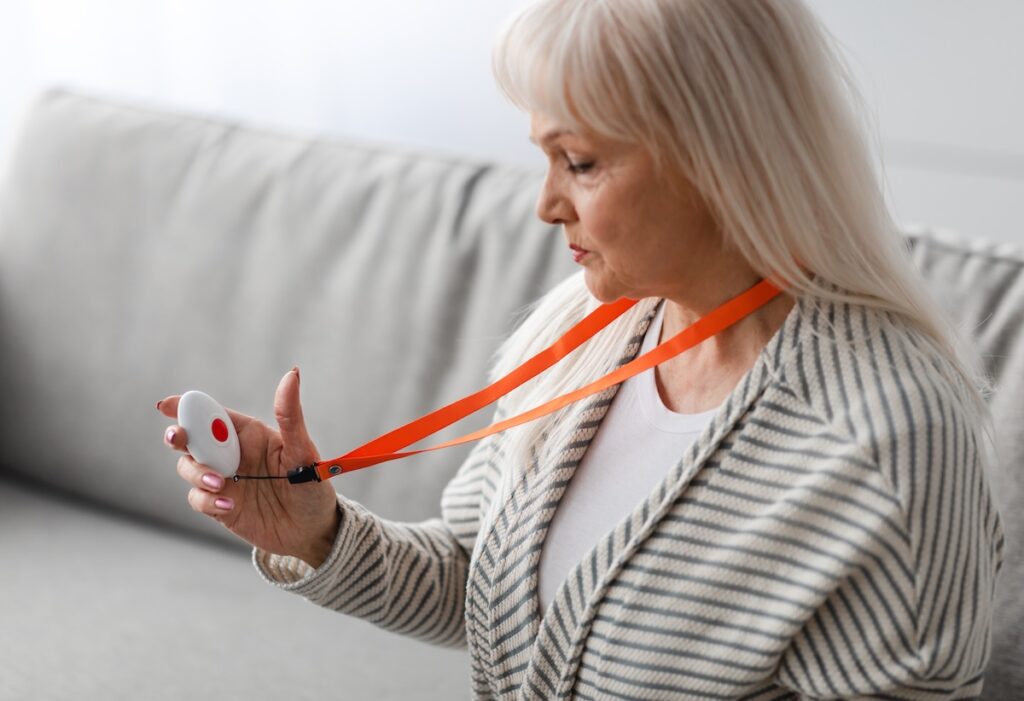 An older woman holds a medical alert button on her hand. It is attached to a red lanyard around the woman's neck