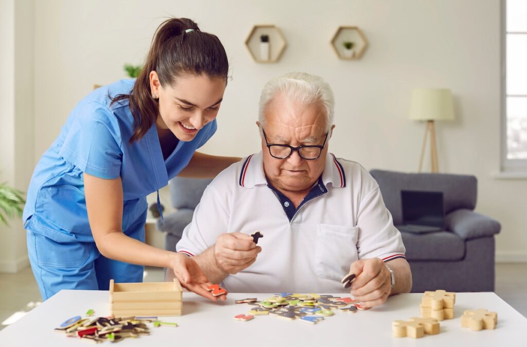 Nurse in geriatric clinic or retirement home helping patient with alphabet puzzle. Senior man sitting at desk and playing games with letters. Old age, dementia, Alzheimer's disease, therapy concept